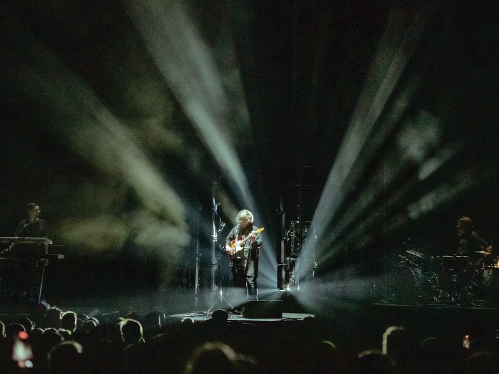 A shot of Alt-J performing on stage from the point of view of an audience member. The main member is playing guitar with back-lit lights behind him.
