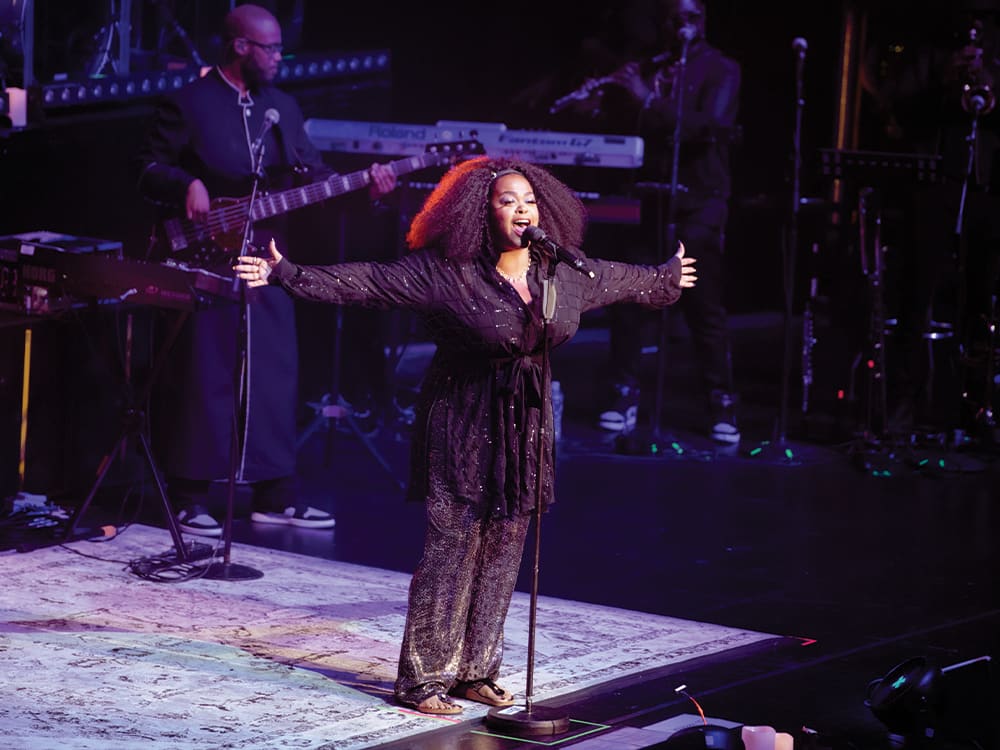 A shot of Jill Scott performing at Kings Theatre. She is a Black woman with dark curly hair. She is smiling with her arms outstretched. She is wearing all black.