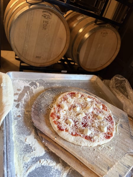 Unbaked pizza topped with sauce, shredded and cubed cheese, set on a floured peel near wine barrels