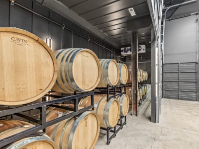 Rows of oak wine barrels on metal racks in a climate-controlled cellar with cooling fans overhead