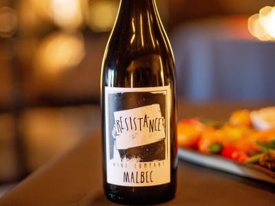 Wine bottle labeled Resistance Malbec on a dark bar, blurred food and lights behind it.