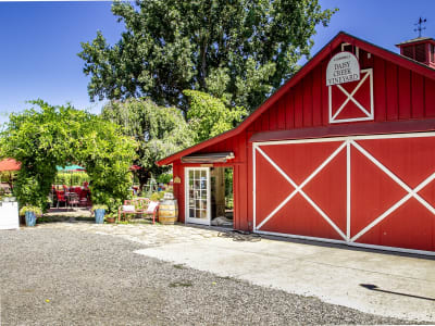 Red barn-style building with white X barn doors and "Daisy Creek Vineyard" sign, patio and trees nearby.