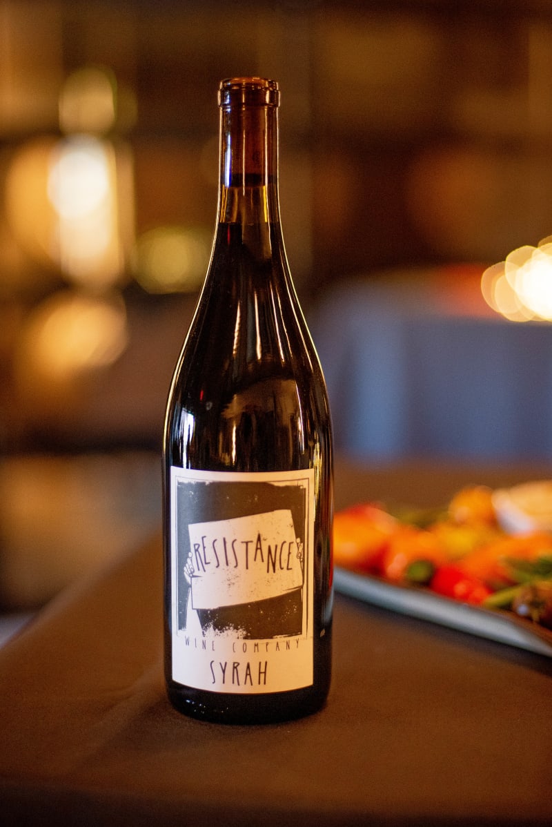 Bottle of Resistance Wine Company Syrah on a table, blurred food and lights behind