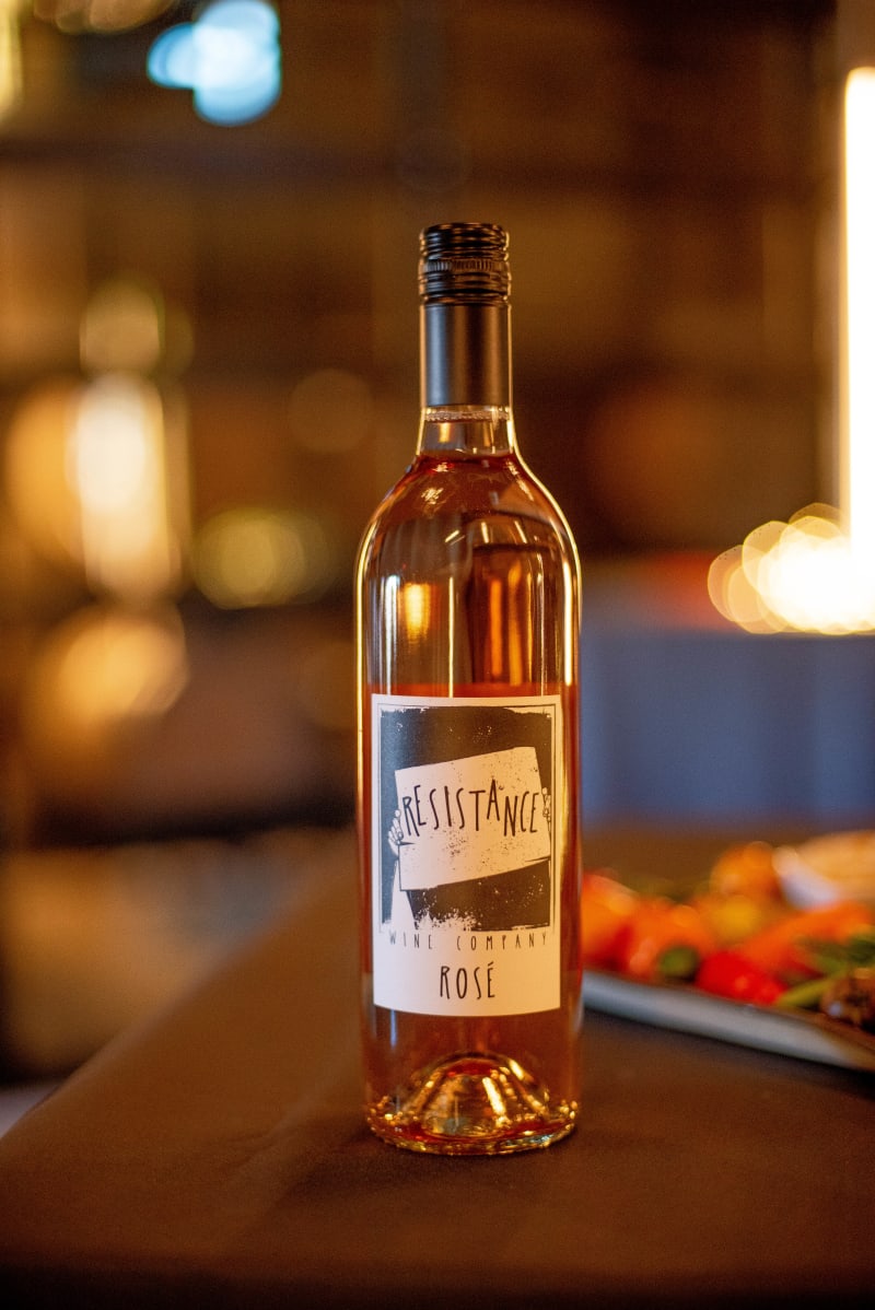 Bottle of Resistance Wine Company Rosé on a wood table, dim string lights in background