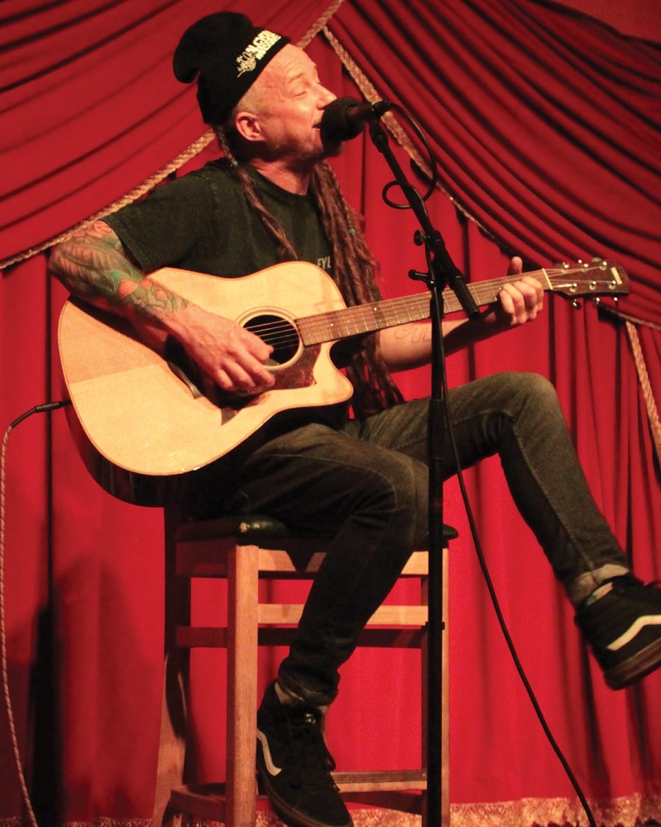 Alcyon Massive's Charlie Blake singing and playing acoustic guitar on a stool in front of a red stage curtain