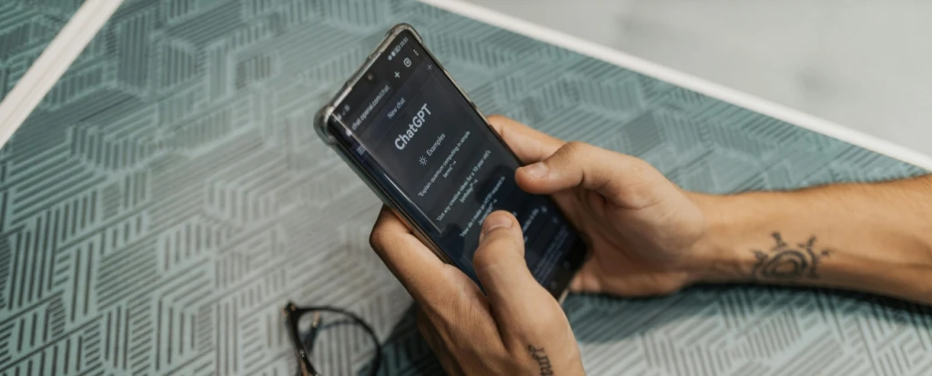 A close-up of hands holding a smartphone displaying a ChatGPT interface. The phone rests on a patterned surface, and a pair of glasses lies nearby.