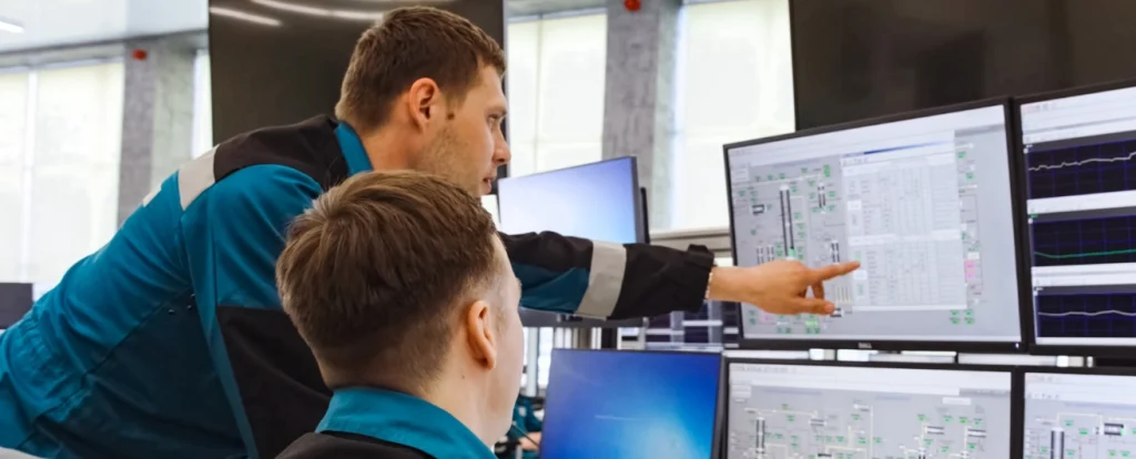 Two technicians in blue uniforms working at a control station with multiple monitors. One technician points at a screen displaying detailed schematics and data, while the other observes attentively. The setting is a modern, well-lit control room.