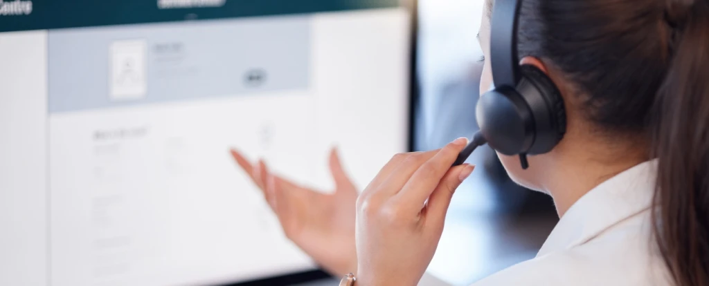 A customer support representative wearing a headset, seated at a desk, interacting with a computer screen displaying a form or user interface. The person's hand is gesturing as they speak, suggesting active communication.