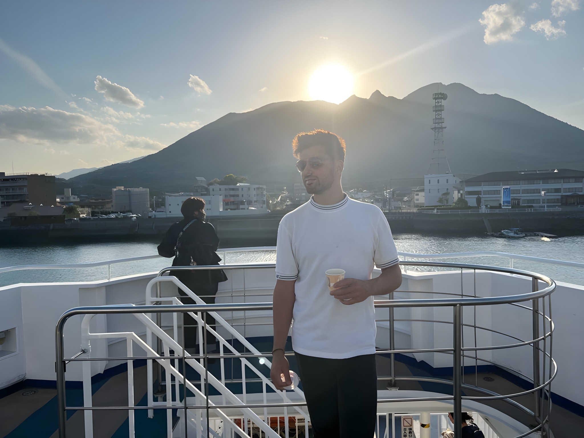A man in a white shirt and sunglasses holds a cup on a ferry deck in Japan during sunset. The warm glow of the sun sets behind a mountain, illuminating budget-friendly cityscapes that inspire ways to save money in Japan. Another individual stands by the railing, gazing out at buildings that promise affordable adventures amidst this scenic voyage.