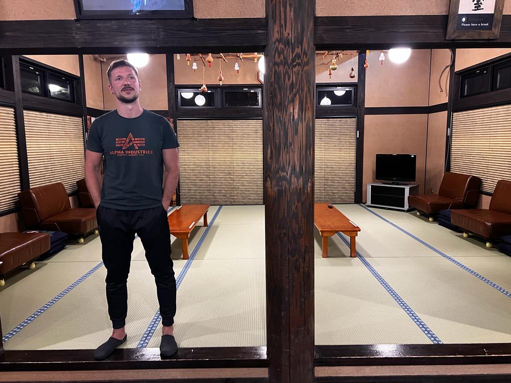 A person relaxes in a traditional Japanese room featuring tatami mats, wooden beams, and wall seating. A TV in the corner offers a chance to save money in Japan by enjoying cost-free entertainment after discovering cheap local eateries and affordable attractions throughout the day. This setting perfectly blends cultural ambiance with budget-conscious travel.