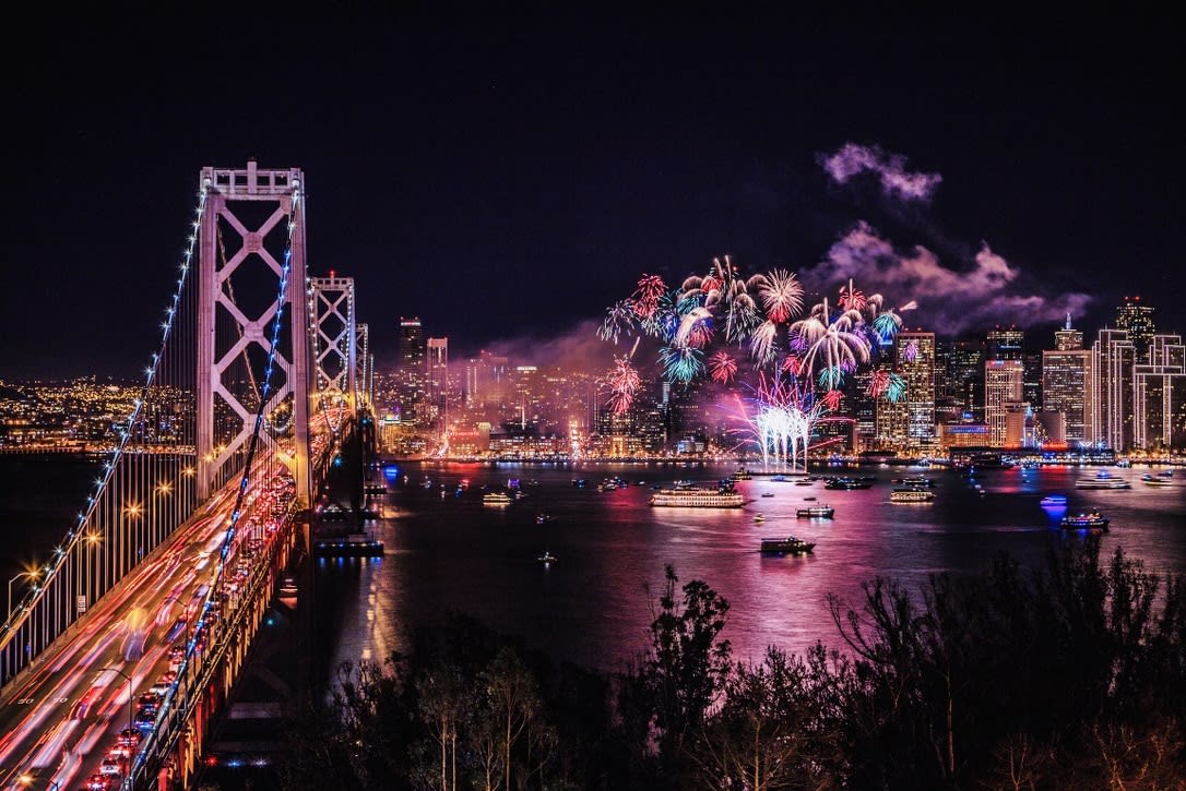 Fireworks light up the night sky over a city skyline and bridge, reflecting on the water below as NYE 2024/2025 celebrations ignite, casting vibrant colors on popular LGBTQ+ hotspots. The energy is electric as people gather for a spectacular gay New Year's Eve, where inclusivity and joy blend with colorful displays and festive spirits.