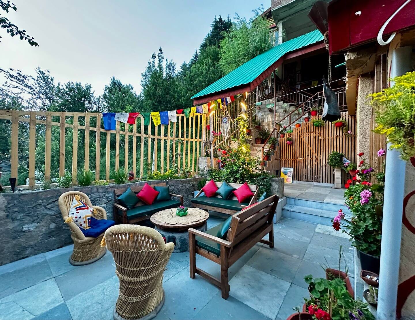 The courtyard: cane chairs and cushioned wooden sofas around a low table on a stone terrace, with prayer flags, flowers and trees