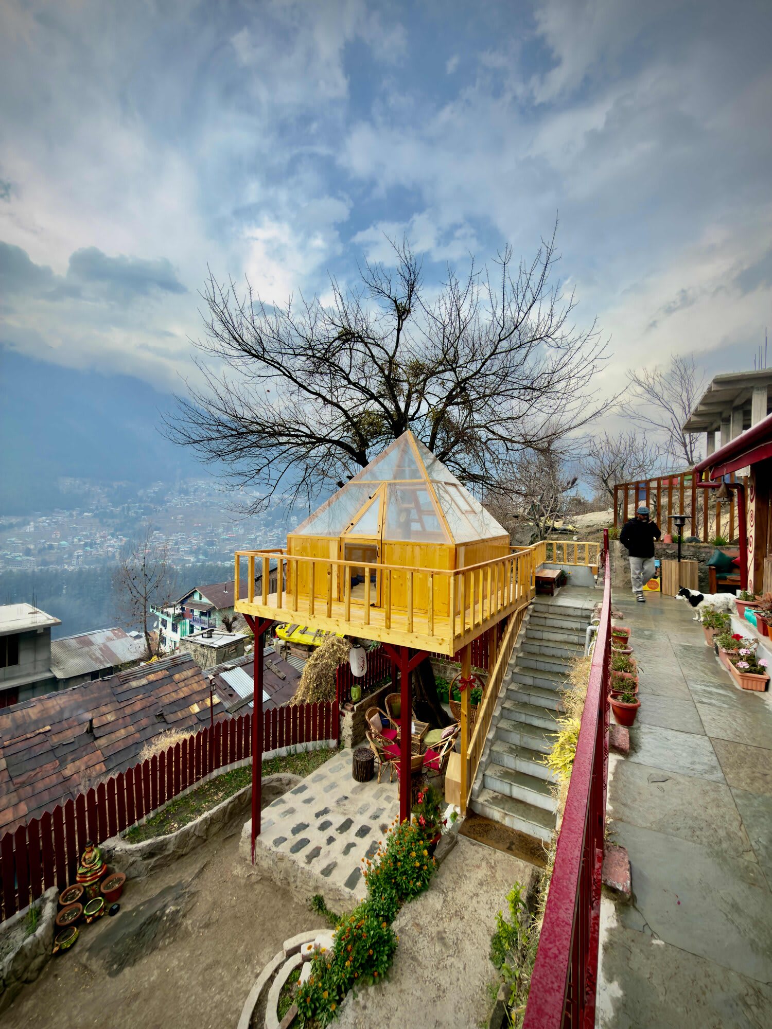 A wooden treehouse with a glass pyramid roof beside stone steps in the garden, with the valley and mountains beyond