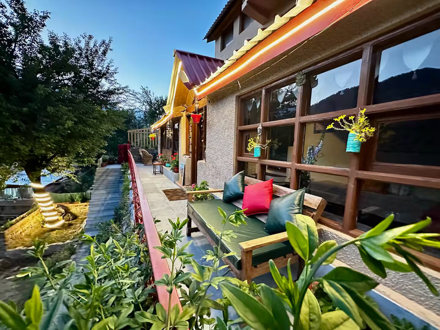 The veranda at dusk: a wooden bench with cushions beside a long row of windows, with plants and a lit railing