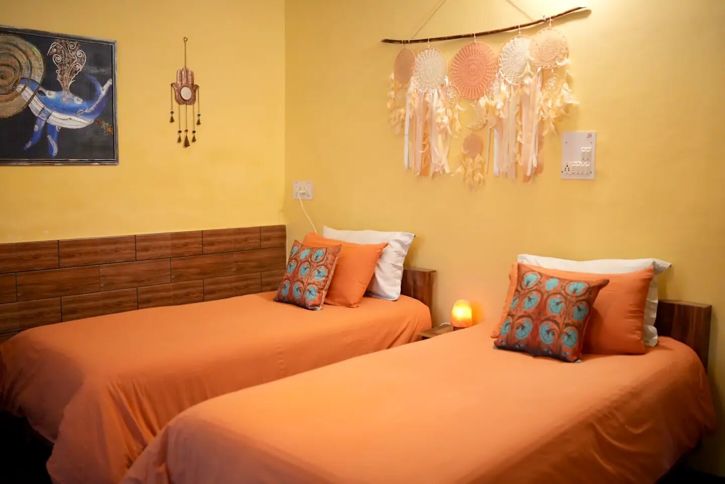 Family Hub: two single beds with orange covers and patterned cushions against a yellow wall hung with dreamcatchers and a painting