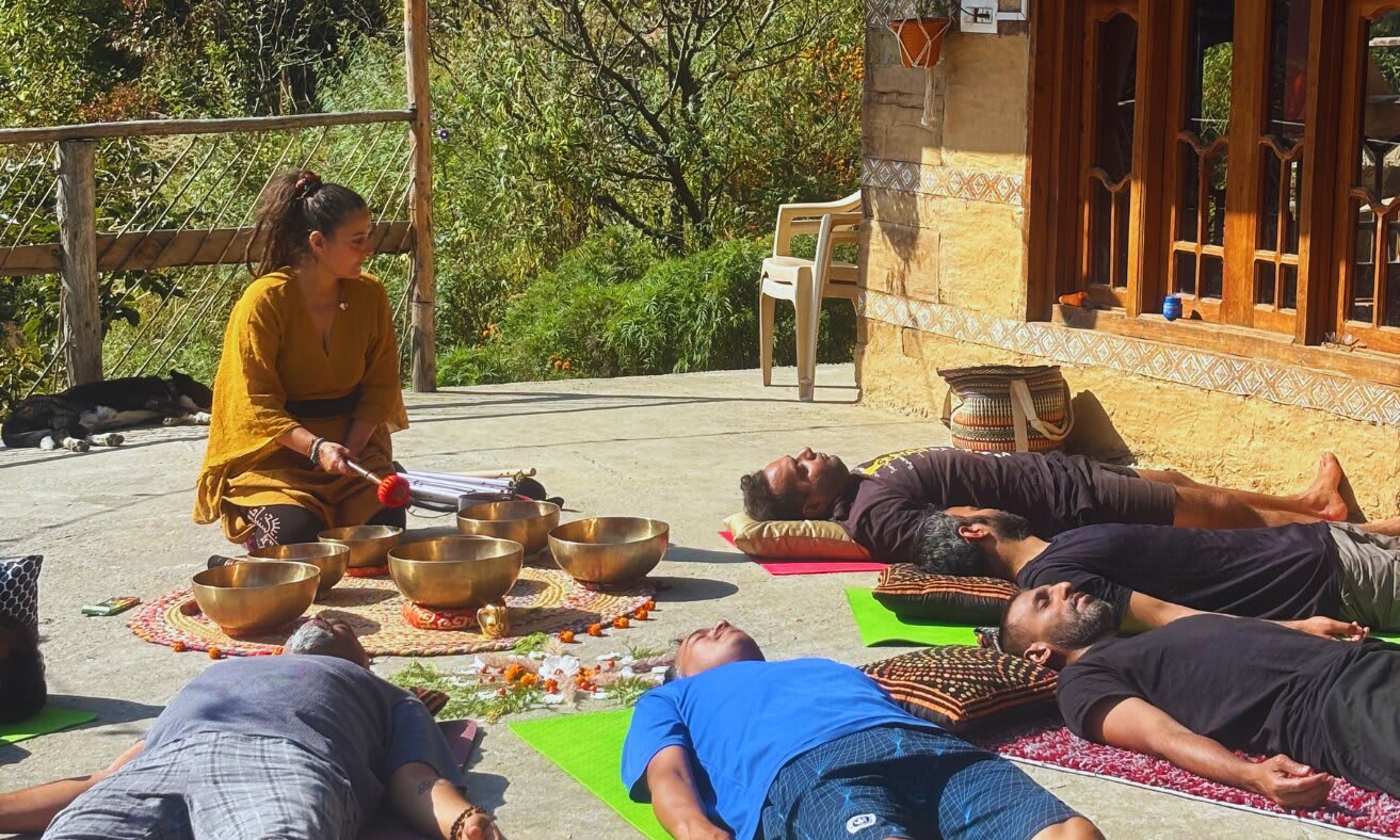 Participants lie on mats on a sunny terrace while a facilitator plays singing bowls