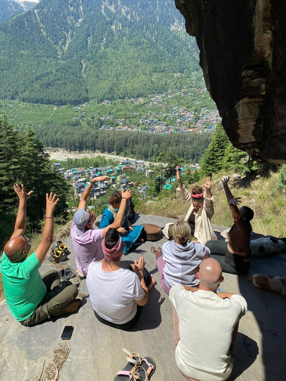 A group sits on a rocky ledge high above the Manali valley, arms raised to the sky