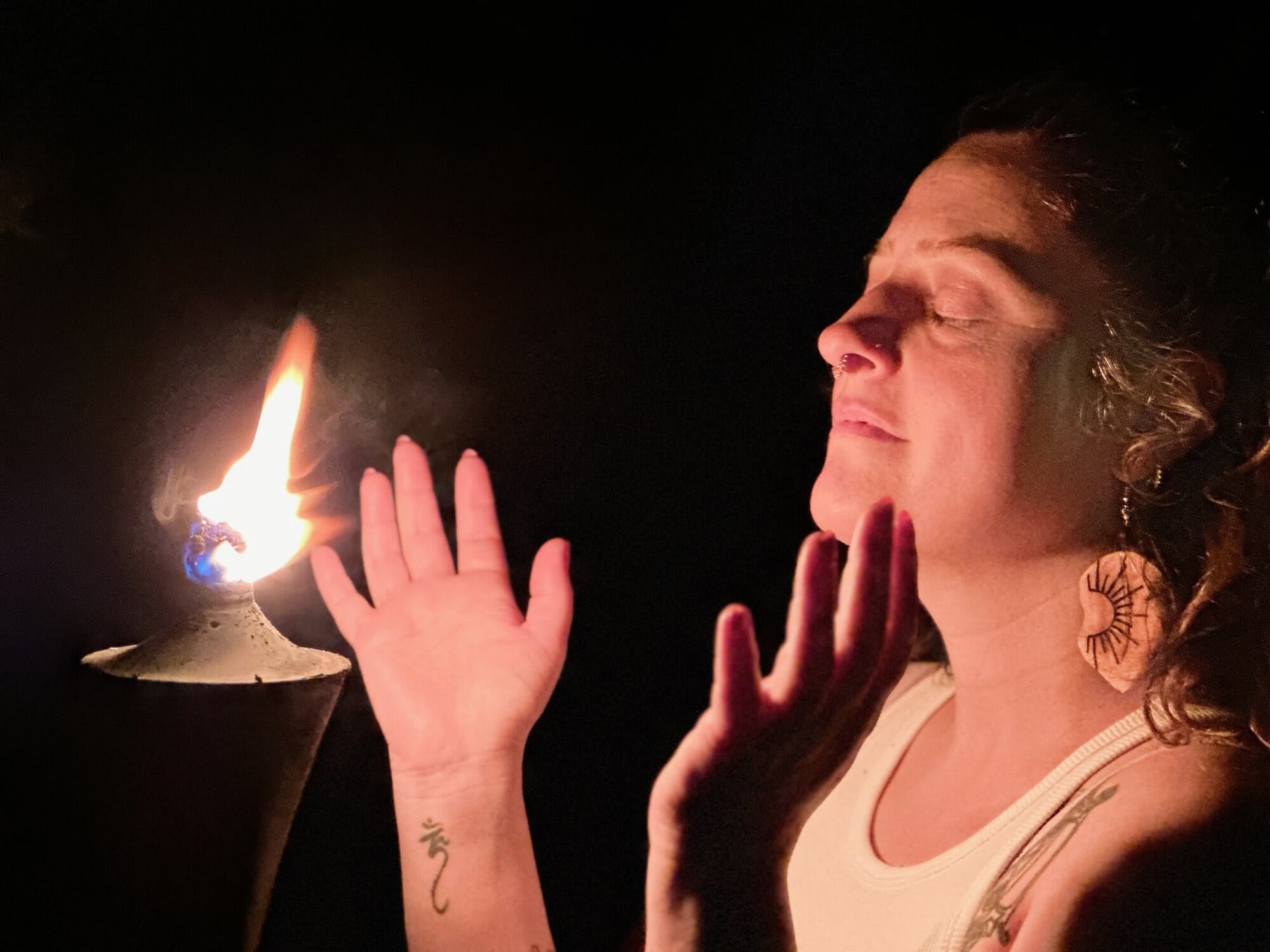 A woman with her eyes closed holds her hands up to a flame in the dark