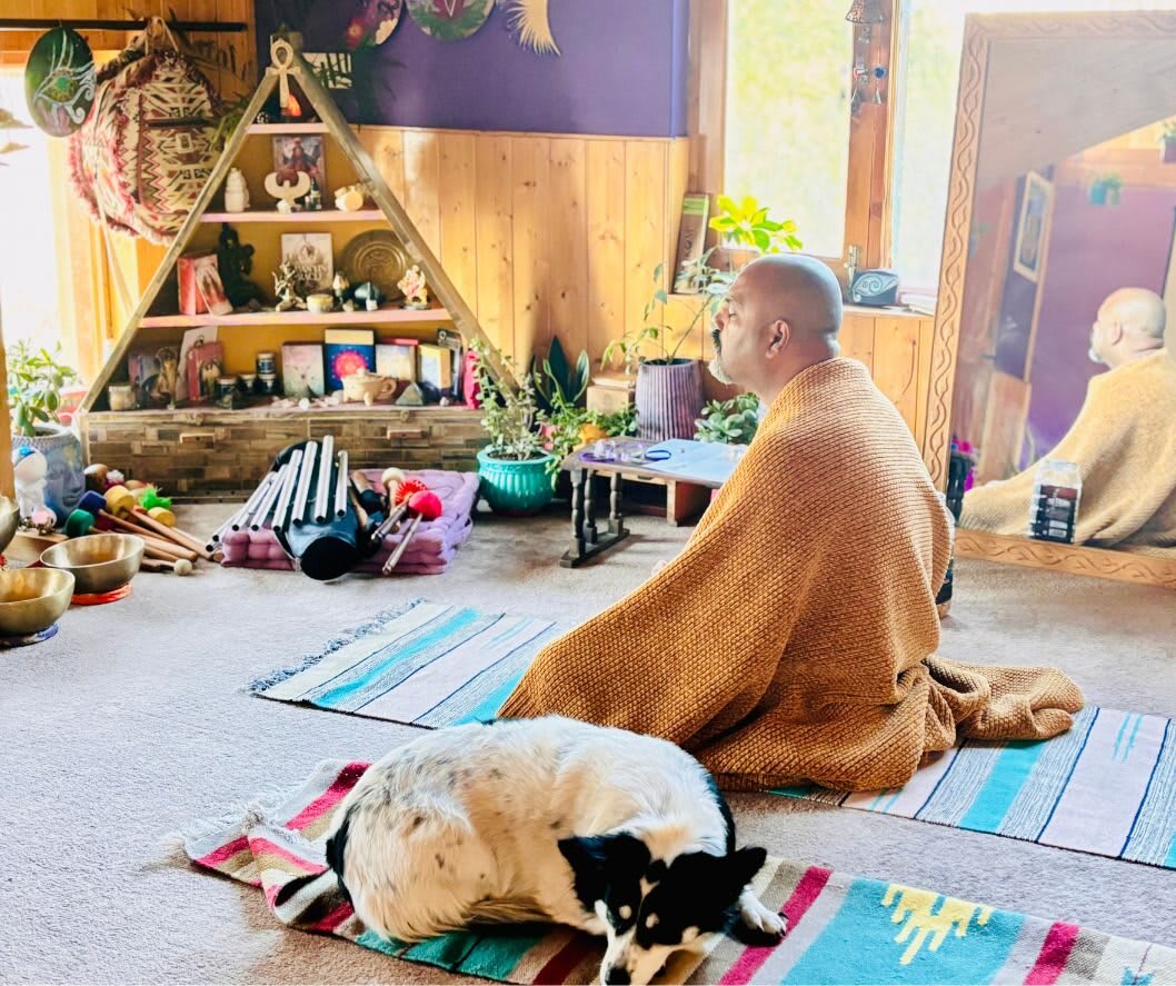 A man wrapped in a shawl meditates on a floor mat in the sacred space at Sehar, a dog sleeping beside him
