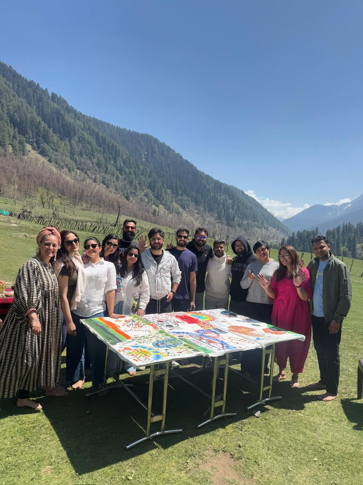 A large retreat group stands beside a table of paintings in a green Himalayan valley