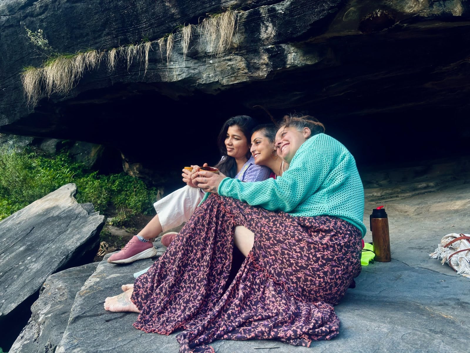 Three women sit together on a rock under an overhanging cliff, smiling and sharing a hot drink