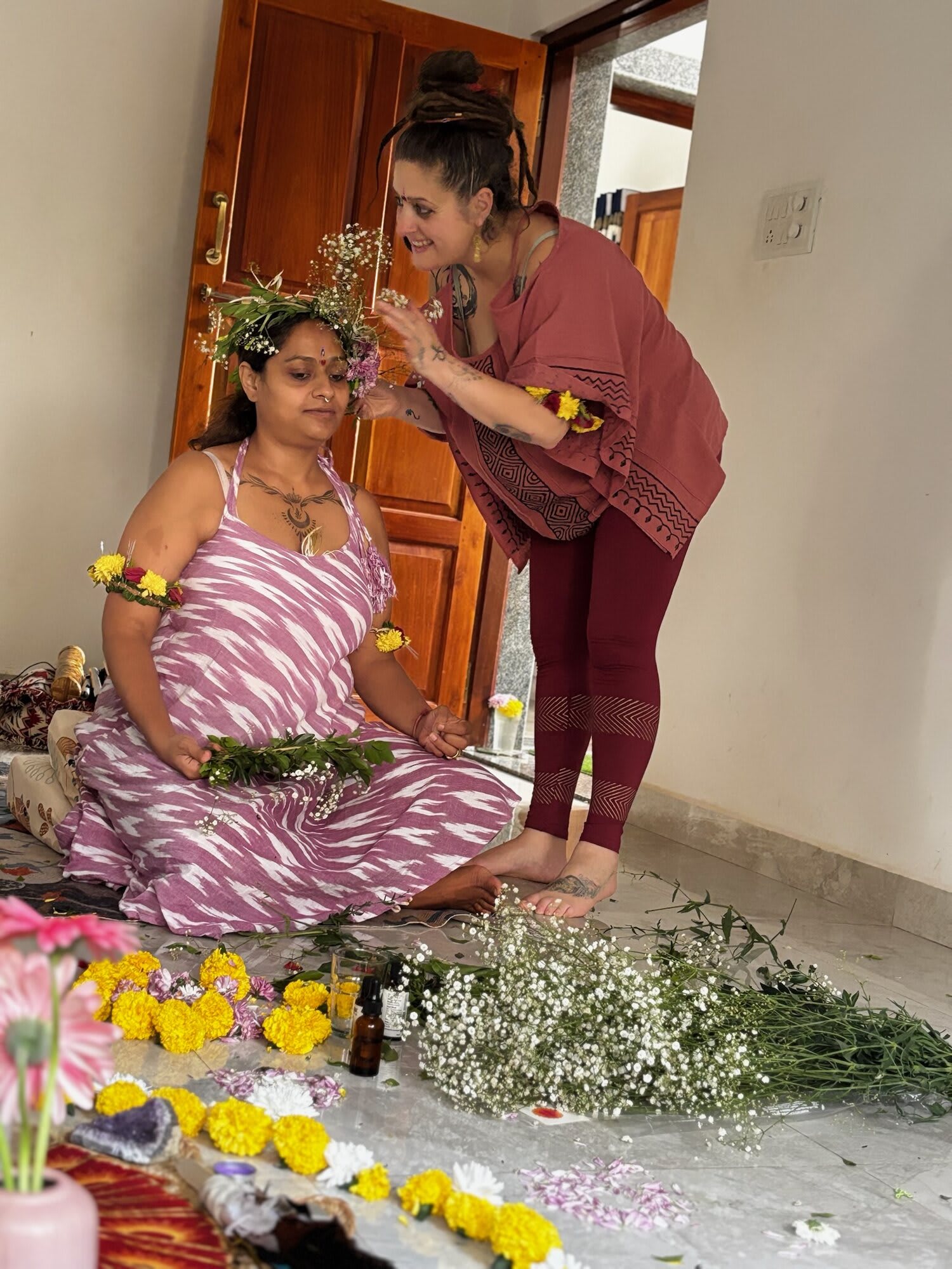 A woman arranges a flower crown on a seated woman in a striped sari, with yellow flowers and greenery at her feet