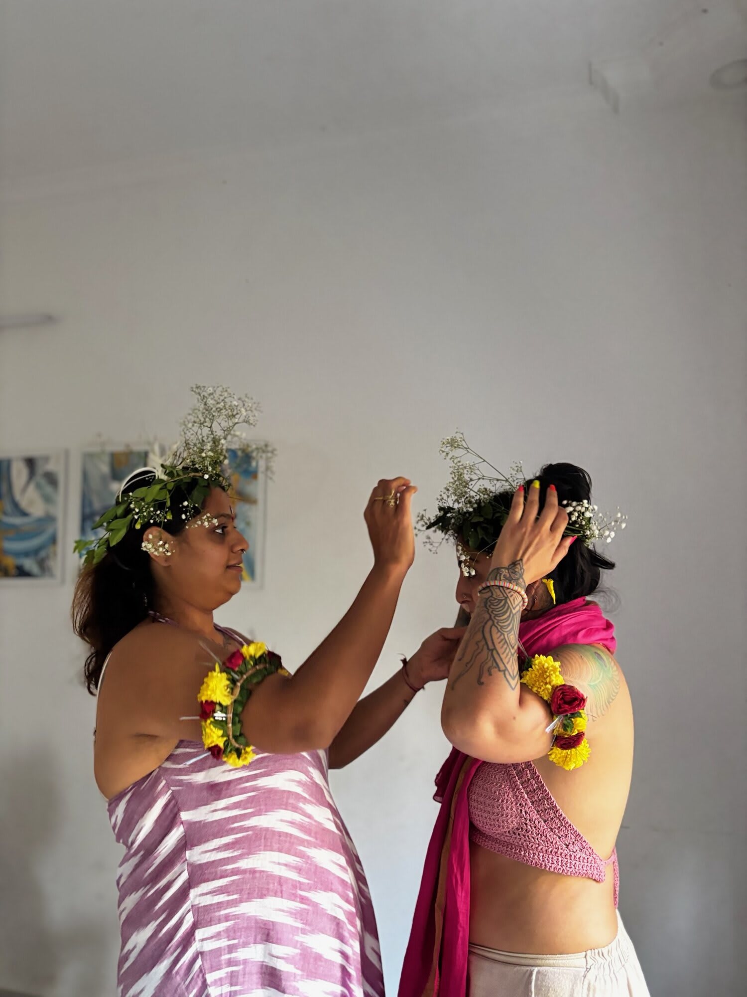 One woman places a flower crown on another's head; both wear garlands of marigolds