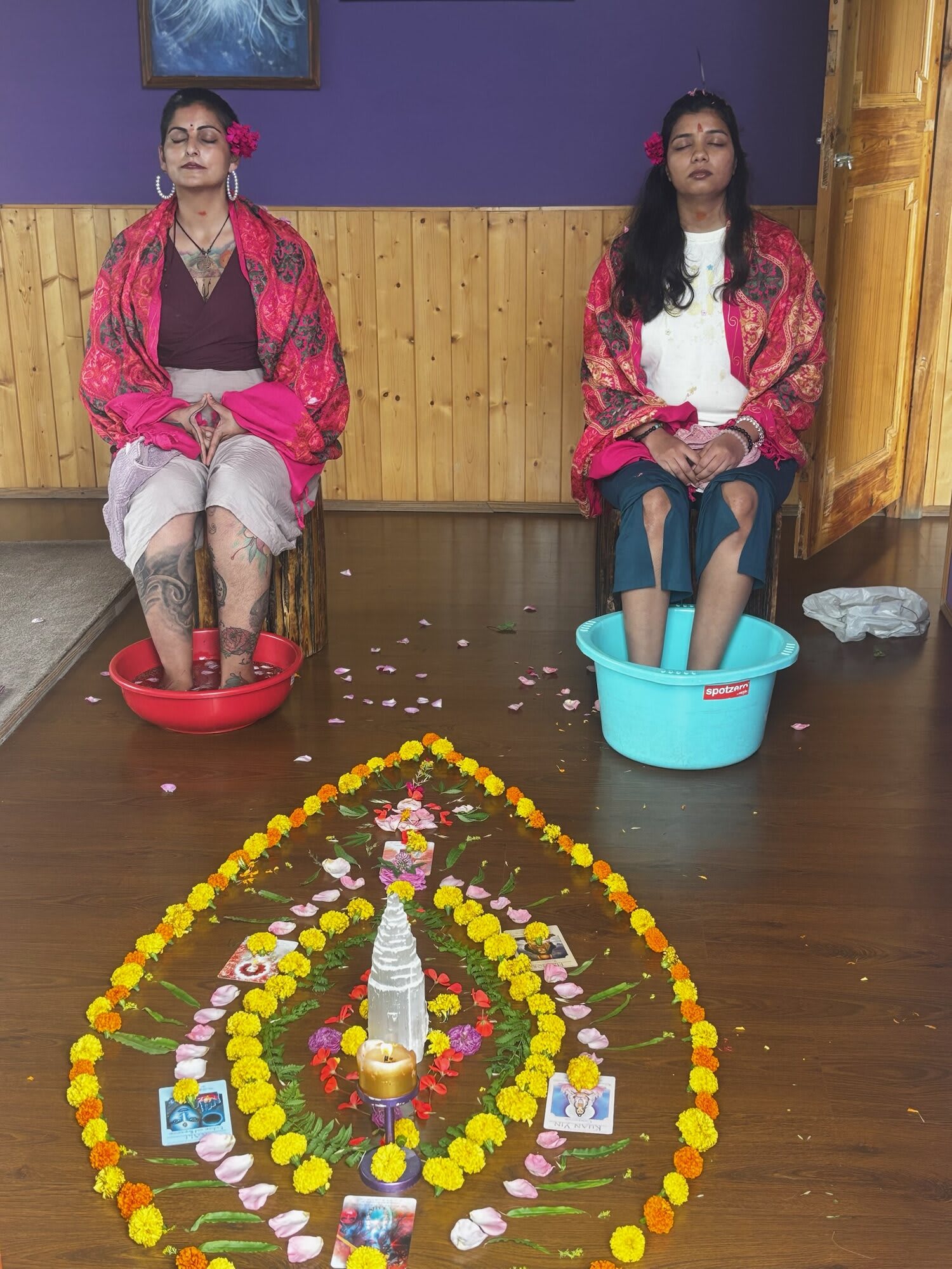 Two women in pink shawls sit with their feet in basins of water behind a flower mandala of marigolds and petals