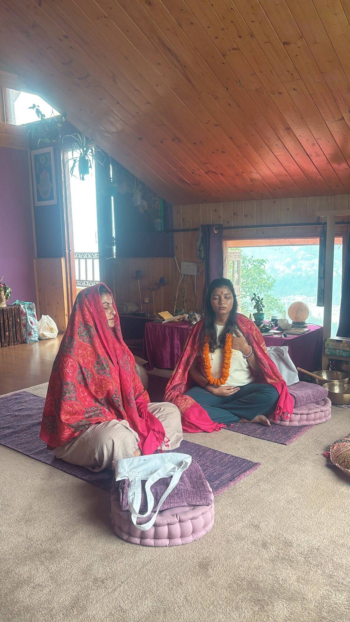 Two women in red shawls sit cross-legged on cushions in a sunlit wooden hall with the mountains outside
