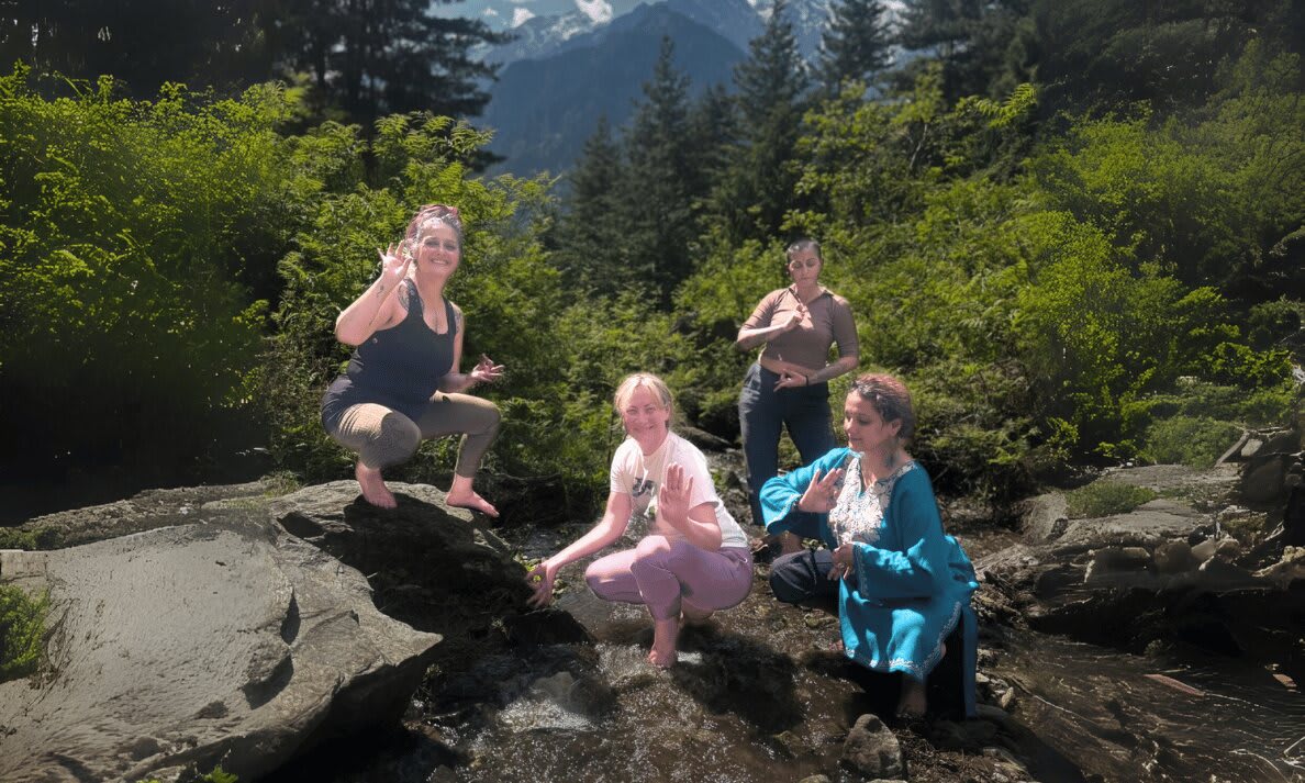 Four women on large boulders in a pine forest, with snow-capped peaks behind