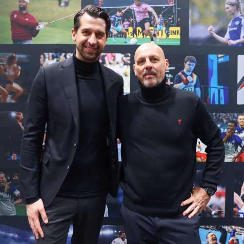 Zwei Männer in dunkler Kleidung stehen vor einer Wand mit Sportbildern.