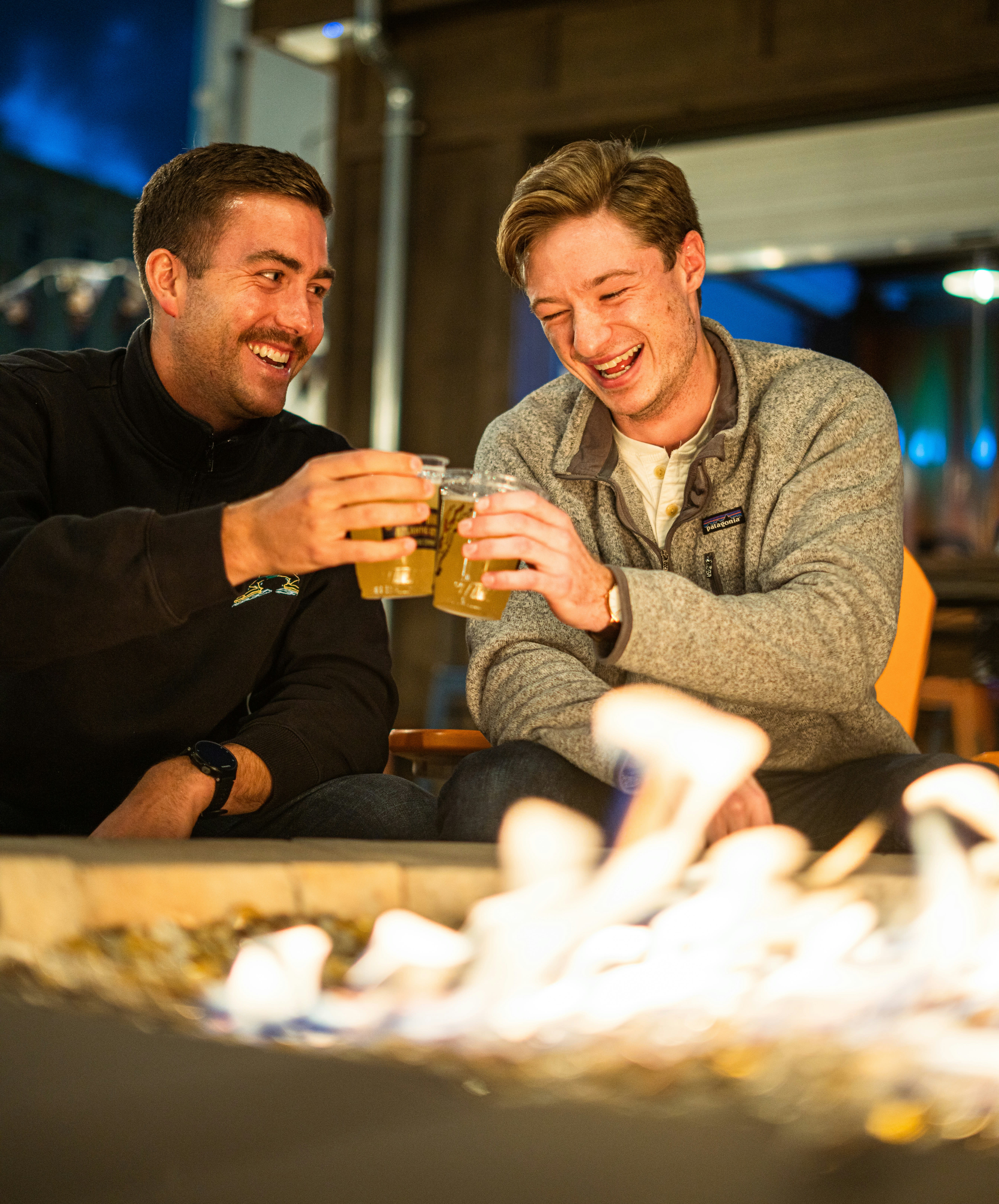 Two friends toasting with beers by a fire pit at night