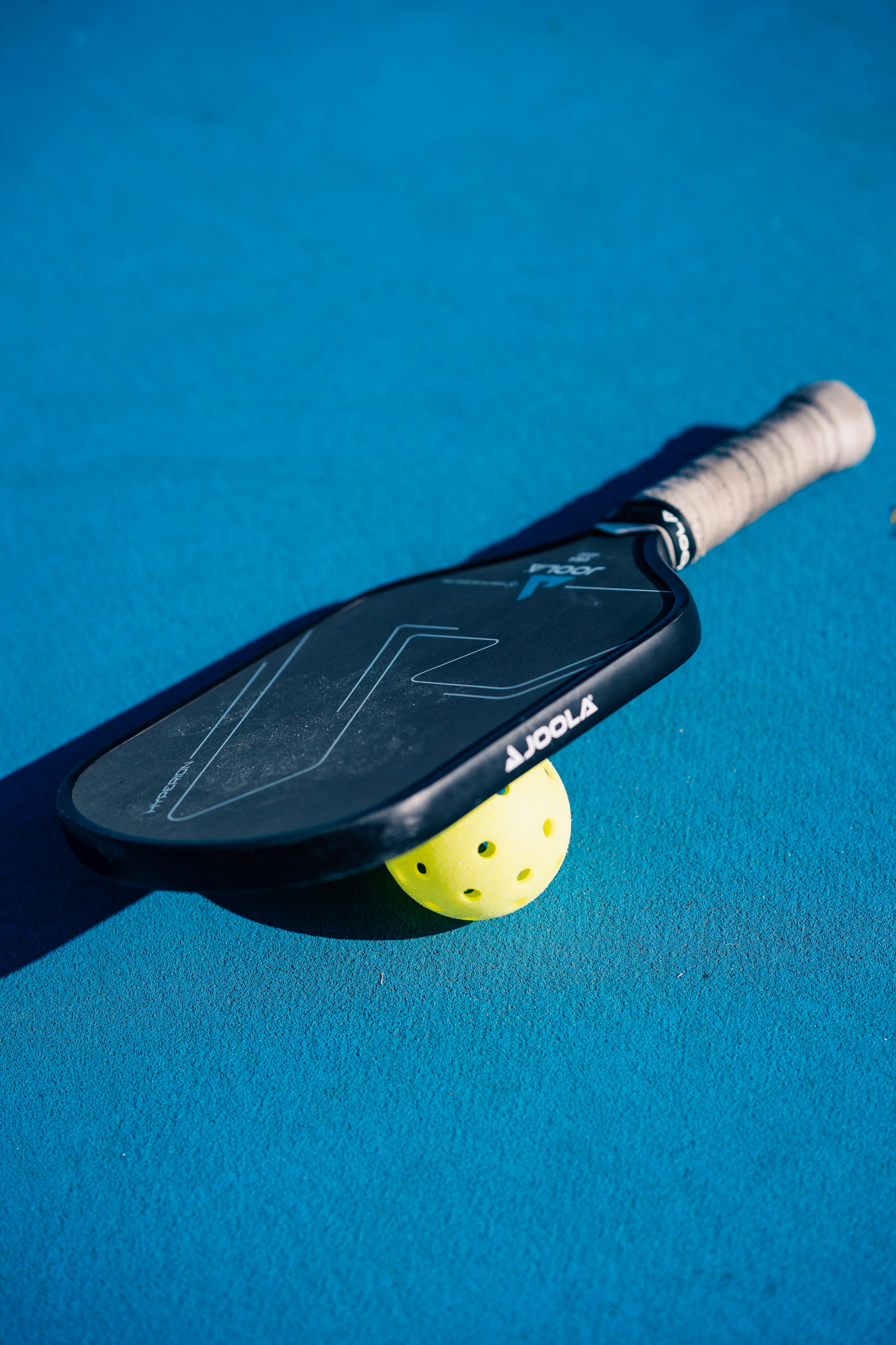 JOOLA pickleball paddle resting beside a neon ball on a vivid blue court surface