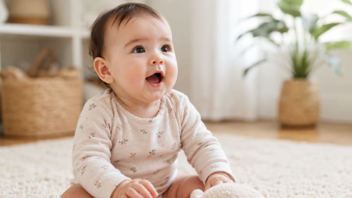 Aydınlık ve sıcak bir ev ortamında otururken ilk seslerini çıkarmaya çalışan, meraklı bakışlara sahip 6–9 aylık bebek.