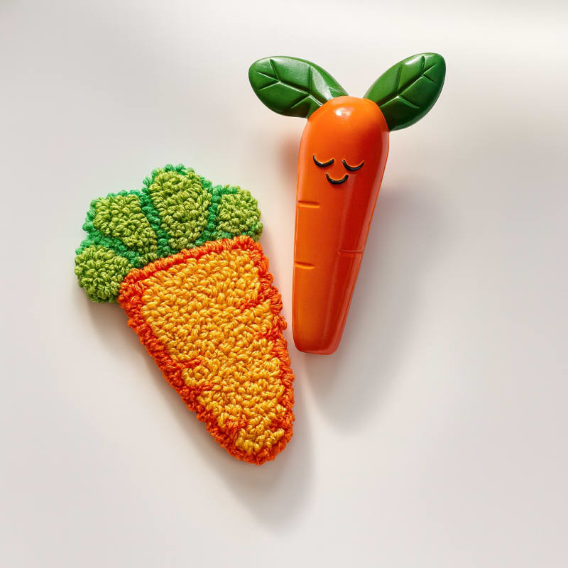 Оранжевый детский прорезыватель в форме моркови с милым личиком в руке. На заднем плане видна коробка продукта и цветы.