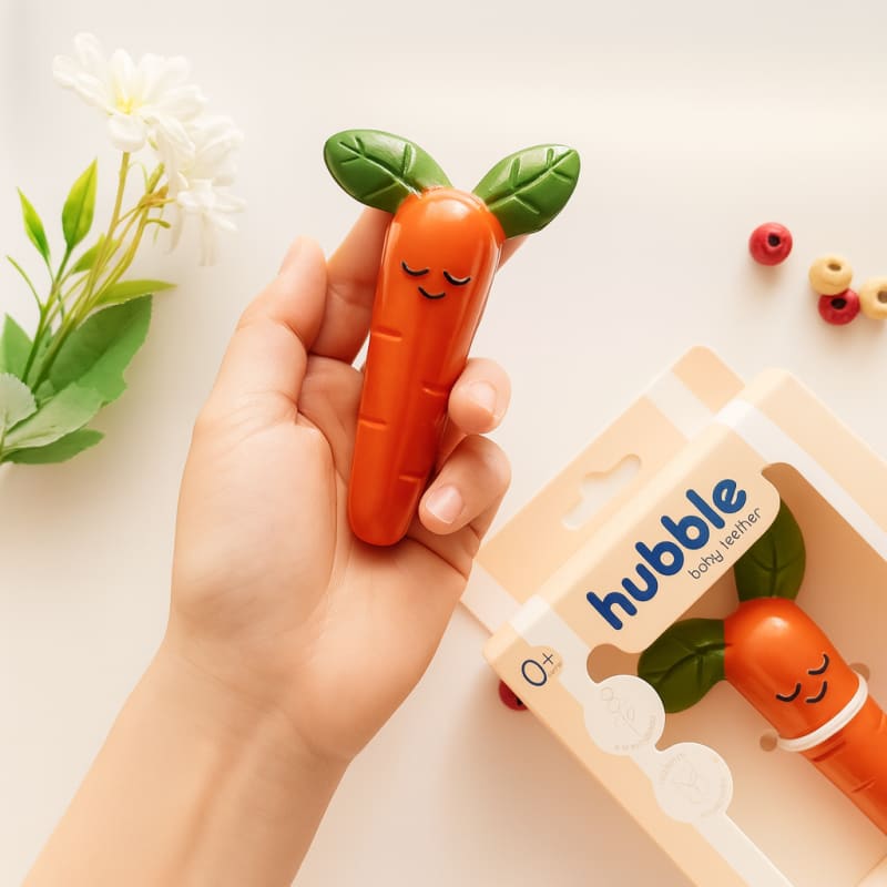 Прорезыватель в форме моркови от бренда Hubble показан рядом с упаковкой, на которой написано "100% натуральный каучук".