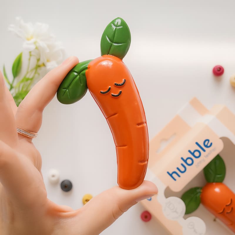 На белом фоне лежит прорезыватель в форме моркови с милым личиком, а рядом с ним - другой текстурированный предмет в форме моркови.