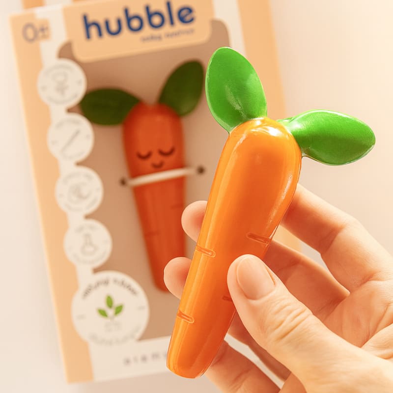 Рука держит детский прорезыватель Hubble в форме моркови. Рядом лежит упаковка продукта и белые цветы.