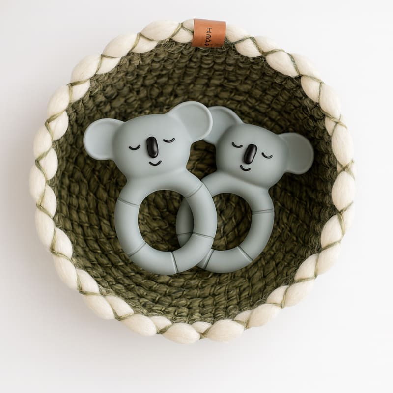 Детский прорезыватель Hubble "Коала" в руке на фоне фирменной коробки и зеленых растений.