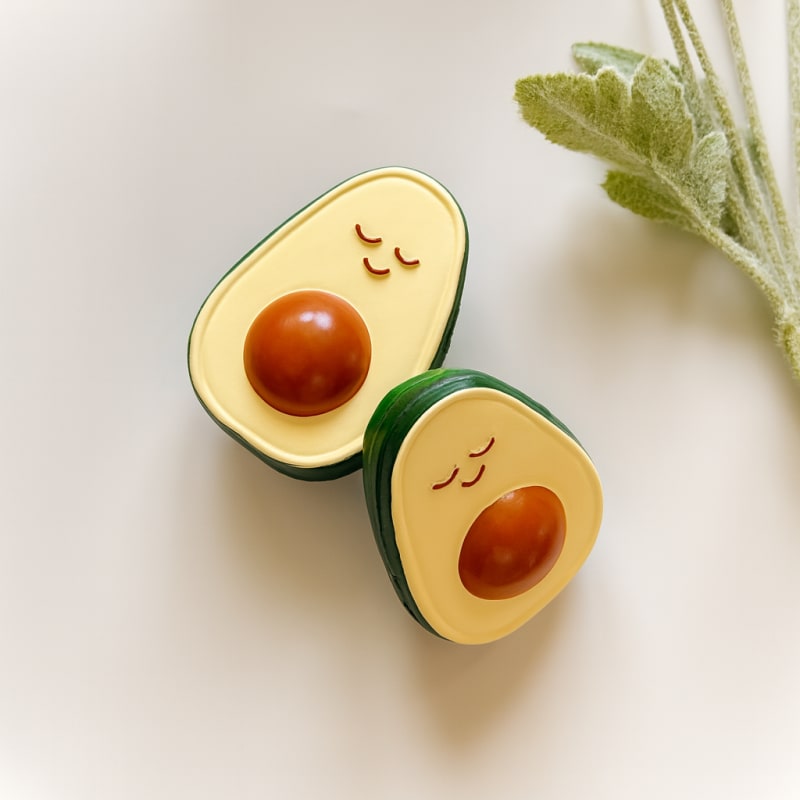 Gülen yüzlü ve çekirdek detaylı, doğal kauçuktan üretilmiş iki adet avokado şeklindeki bebek diş kaşıyıcı; yeşil yaprak dekorlu doğal konsept çekimi.