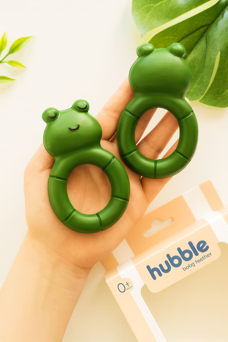 Avuç içinde yan yana sergilenen iki adet sevimli yeşil kurbağa şeklindeki bebek diş kaşıma halkası ve arka planda Hubble markalı ambalaj kutusu.