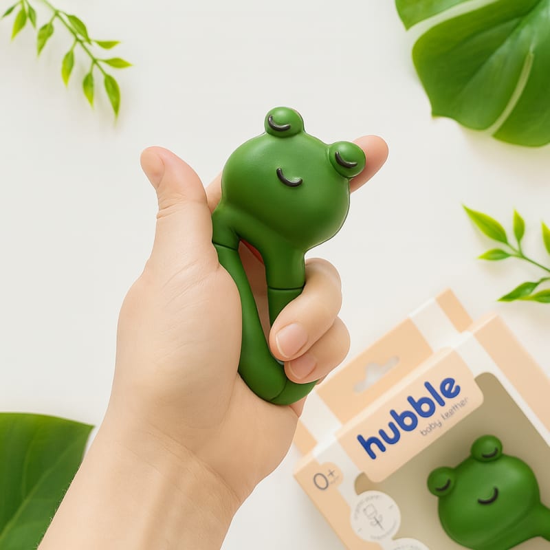 Hubble marka kutusunun yanında, bir elde sıkıca kavranmış yeşil kurbağa figürlü doğal kauçuk bebek diş kaşıyıcı; ergonomik tutuşu gösteren detaylı fotoğraf.