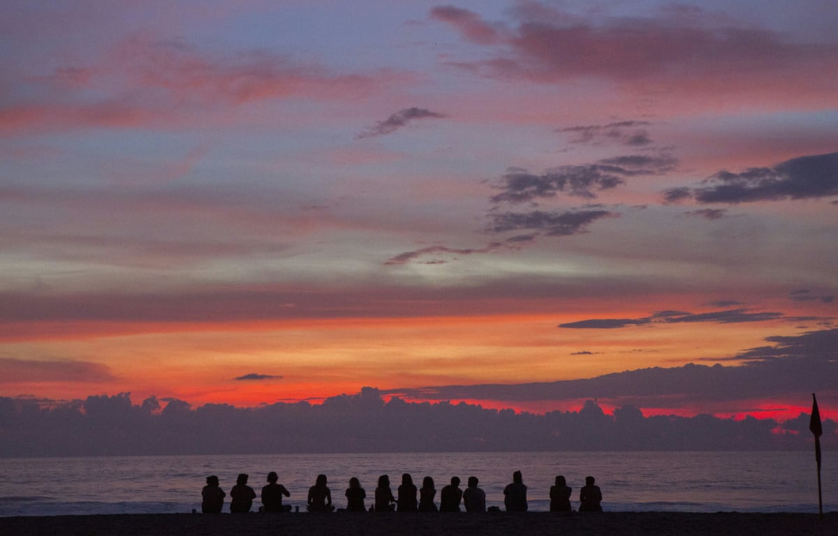People sitting in a row on the sand watching a red and violet sunset over the Pacific, with a red no-swimming flag at the edge of the beach