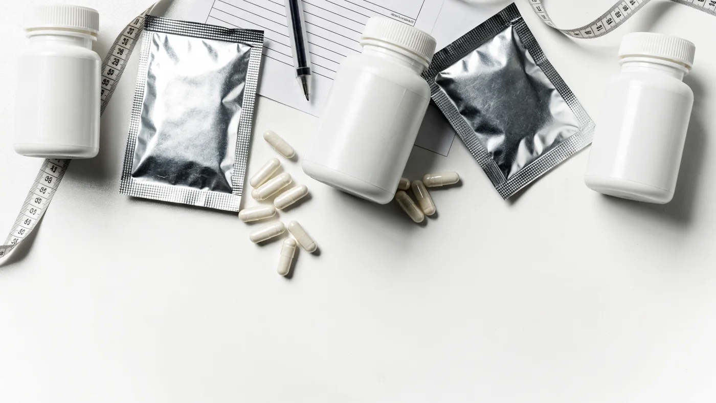 Vista desde arriba de una mesa con muestras de producto: botes sin etiquetar, sachets sellados, cápsulas sueltas, una hoja de especificación con pluma y una cinta de medir