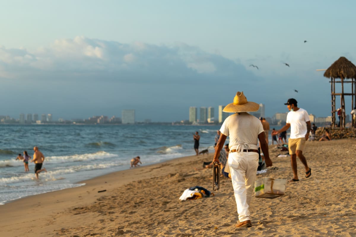 A beach vendor in a straw hat walking along the sand in Puerto Vallarta with the Hotel Zone towers in the distance
