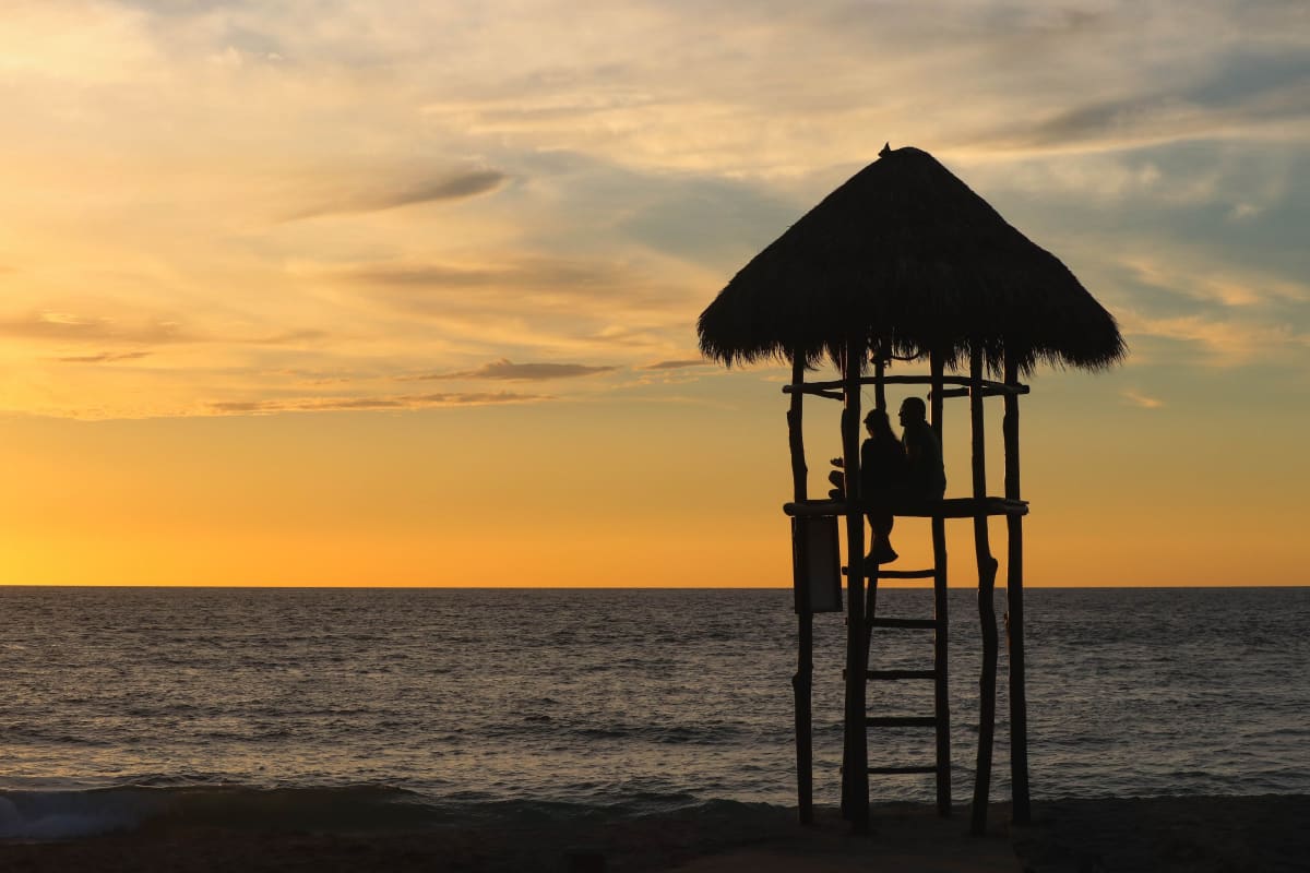 Silhouette of a wooden lifeguard tower with a palm-thatched roof against an orange sunset over the ocean