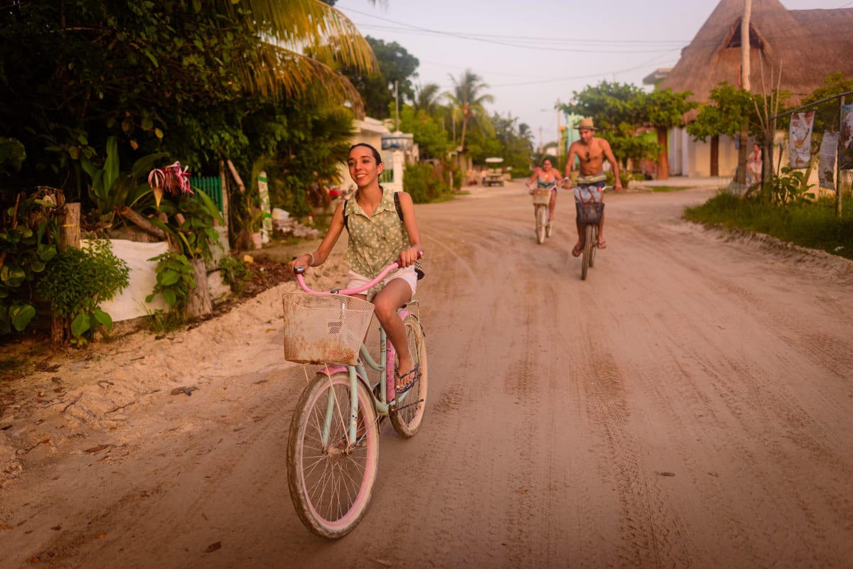 People cycling down a sandy street lined with palm trees and thatched roofs in downtown Holbox, a good area for where to stay in Holbox on foot