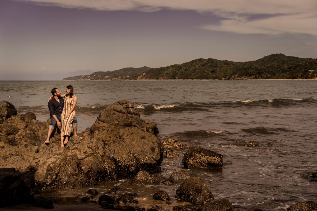 Couple assis sur des rochers sombres au bord de la mer près de Sayulita, collines vertes en arrière-plan
