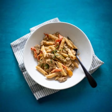 Small batch portion of Rasta pasta with chicken in a white bowl with a black fork on the side over a blue background