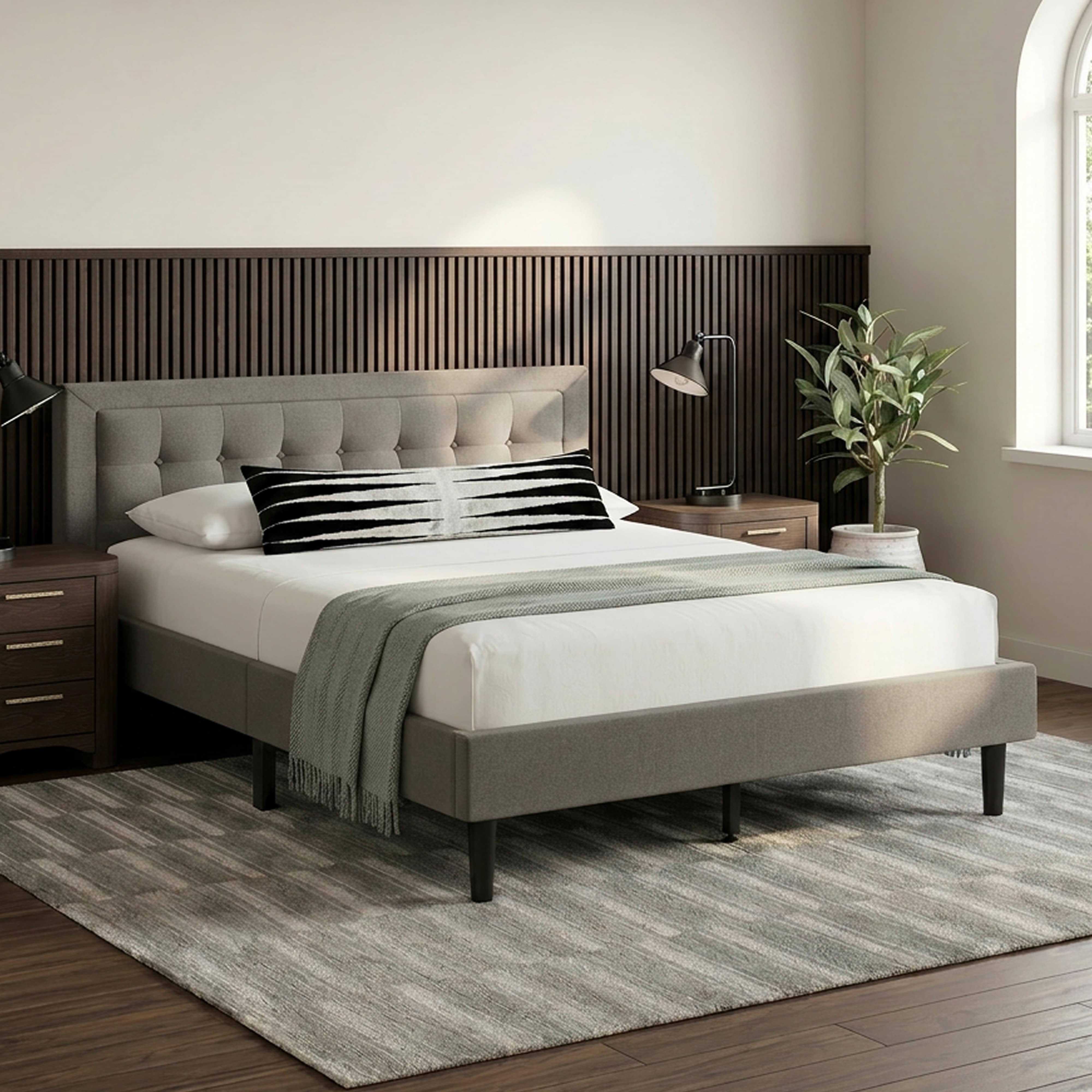 A grey bed with a tufted headboard, white bedding, and a green throw blanket in a bedroom with wood paneling.